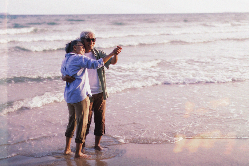 An elderly Asian couple dancing in the tide on the beach, both barefoot with their pants rolled up.