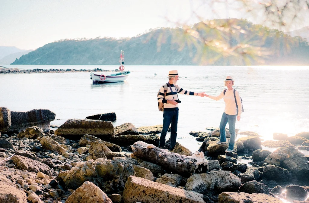 middle age couple holding hands as they walk along a rocky beach with sailboat in the background

